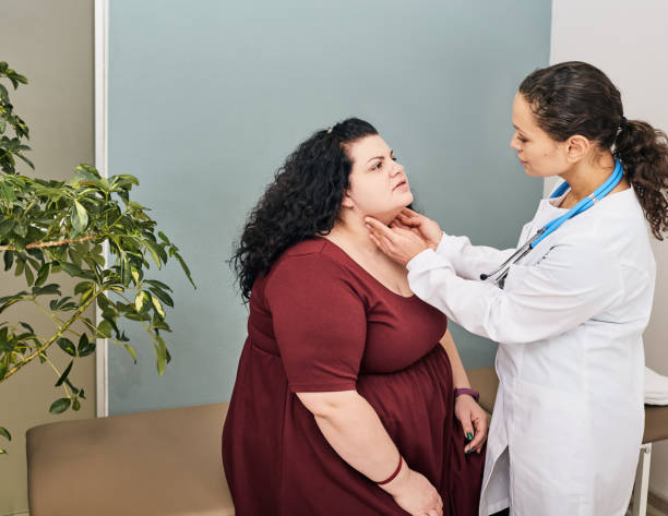 obese woman sitting on the edge of an exam table with a female provider standing in front of her checking her thyroid 
